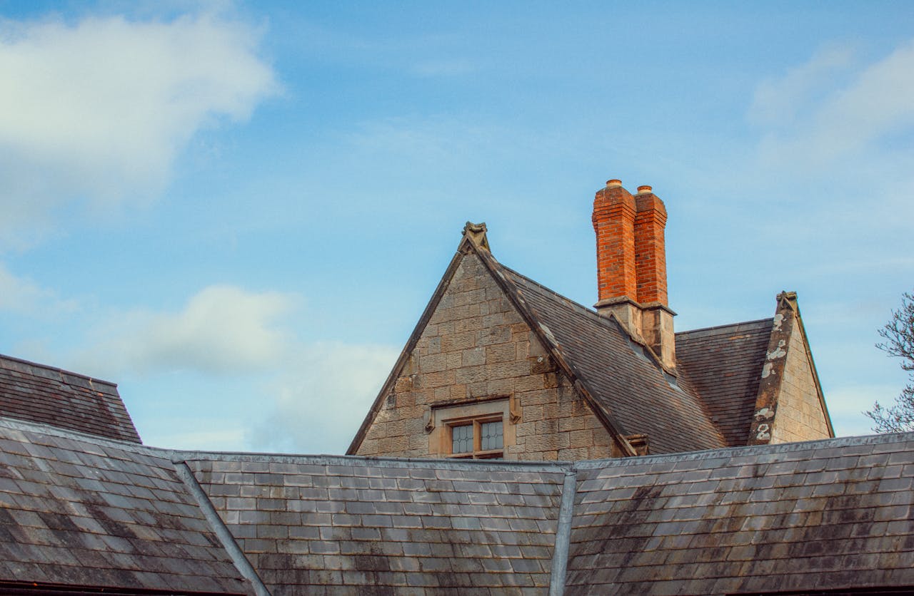 A wide shot of a historic stone building with a slate tile roof and tall brick chimneys, showing natural wear and tear through weathered stone and moss growth, used to contrast with sudden storm damage.