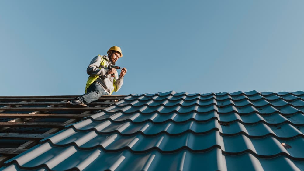A roofer wearing a safety vest and helmet is standing on a blue tiled roof under a clear blue sky, appearing focused and diligent.
